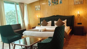 A hotel room with a large bed, two green armchairs, a glass-top table, wooden flooring, and a wooden paneled wall with artwork. - Cottages & Cafe (Kotdwar)