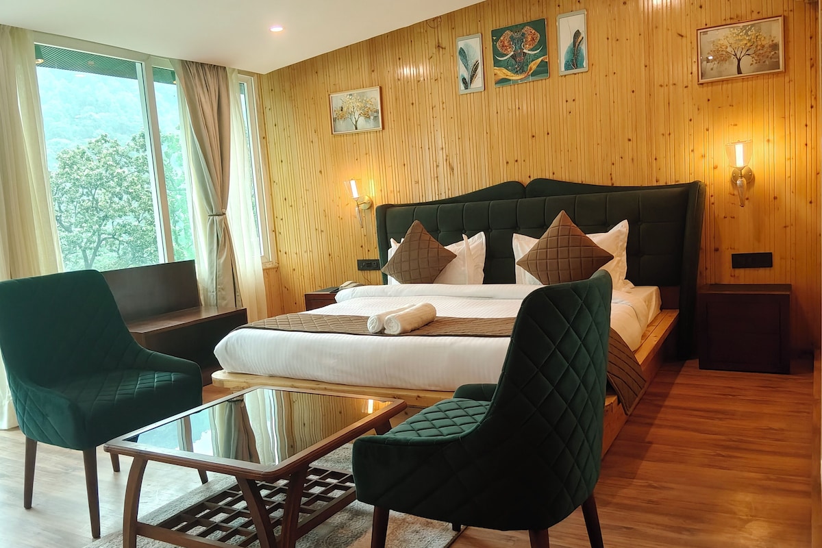 A hotel room with a large bed, two green armchairs, a glass-top table, wooden flooring, and a wooden paneled wall with artwork.