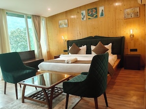 A hotel room with a large bed, two green armchairs, a glass-top table, wooden flooring, and a wooden paneled wall with artwork. - Cottages & Cafe (Kotdwar)