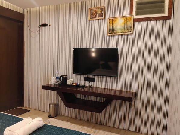 Habitación de hotel con televisor de pantalla plana en la pared, una mesita de madera con hervidor y tazas, un cubo de basura y una toalla enrollada en el suelo.