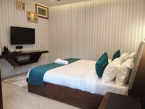 A hotel room with a bed, a flat-screen TV, a wooden side table, and a wall with vertical stripes. - Cottages & Cafe (Kotdwar)