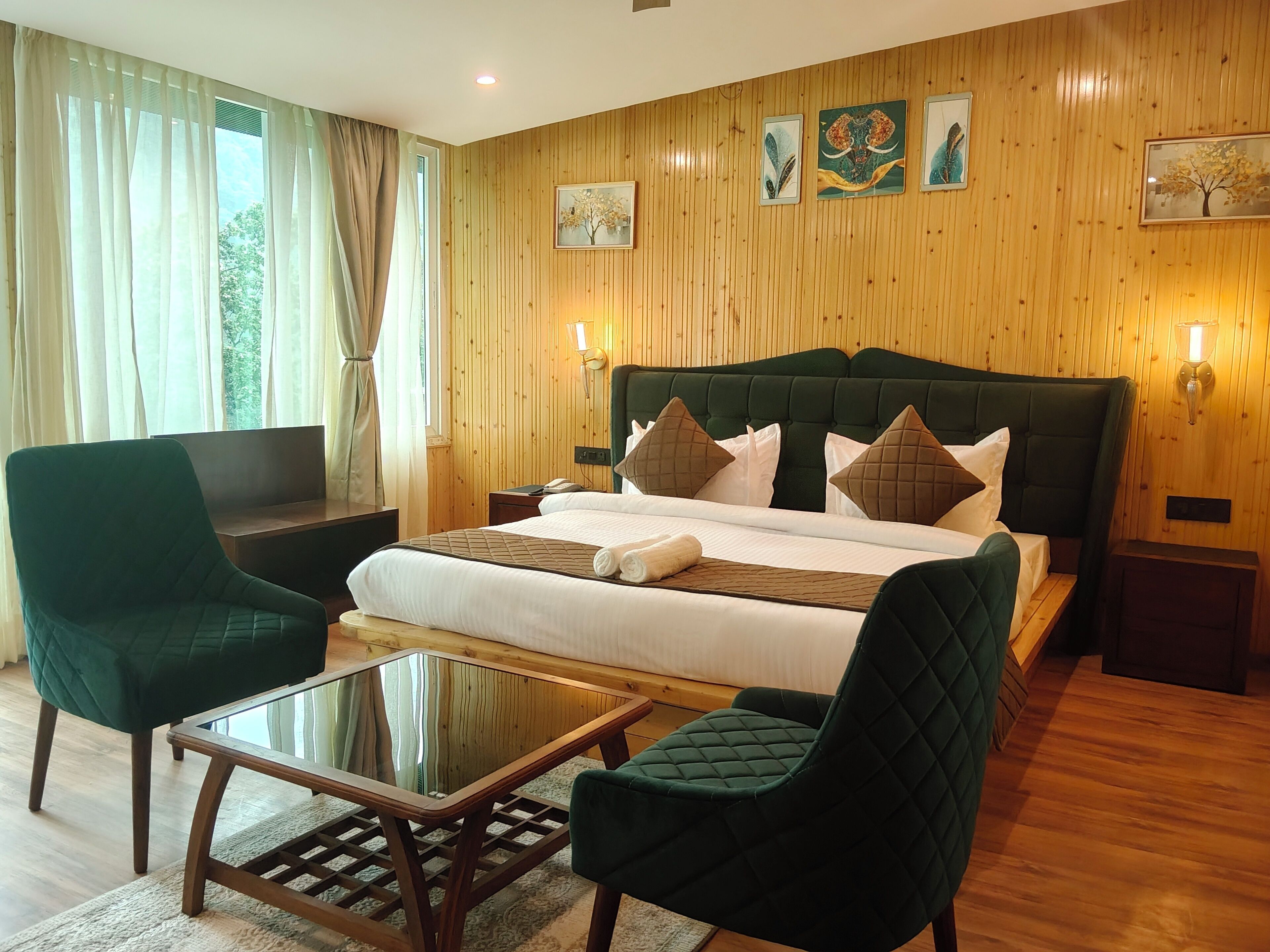 A hotel room with a bed, two green armchairs, a glass-top table, wooden floors, and wooden panel walls.
