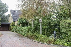 Ein gepflasterter Weg führt zu einem Haus mit Garage, umgeben von üppigem Grün und einer hölzernen Pergola.