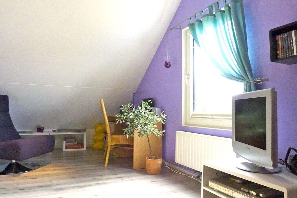 Una habitación con techo inclinado, un televisor sobre un mueble bajo, una planta en maceta y una ventana con cortinas.