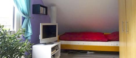 A small room with a bed, a television on a stand, a plant, and a window with curtains.