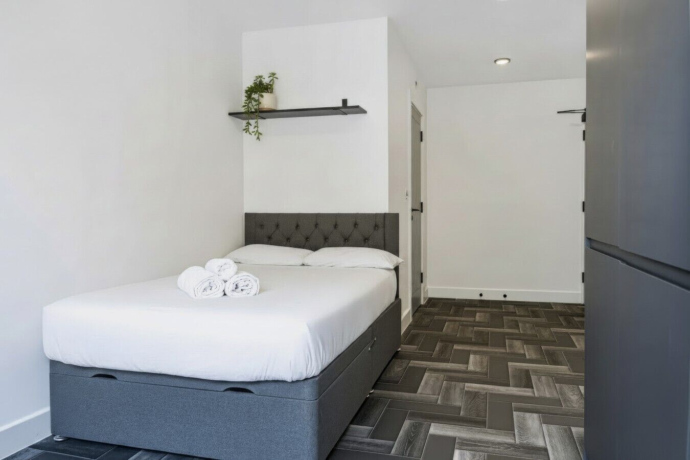 Ein modernes Hotelzimmer mit einem Bett, einer kleinen Ablagefläche mit einer Pflanze und einem Parkettboden mit Fischgrätmuster.