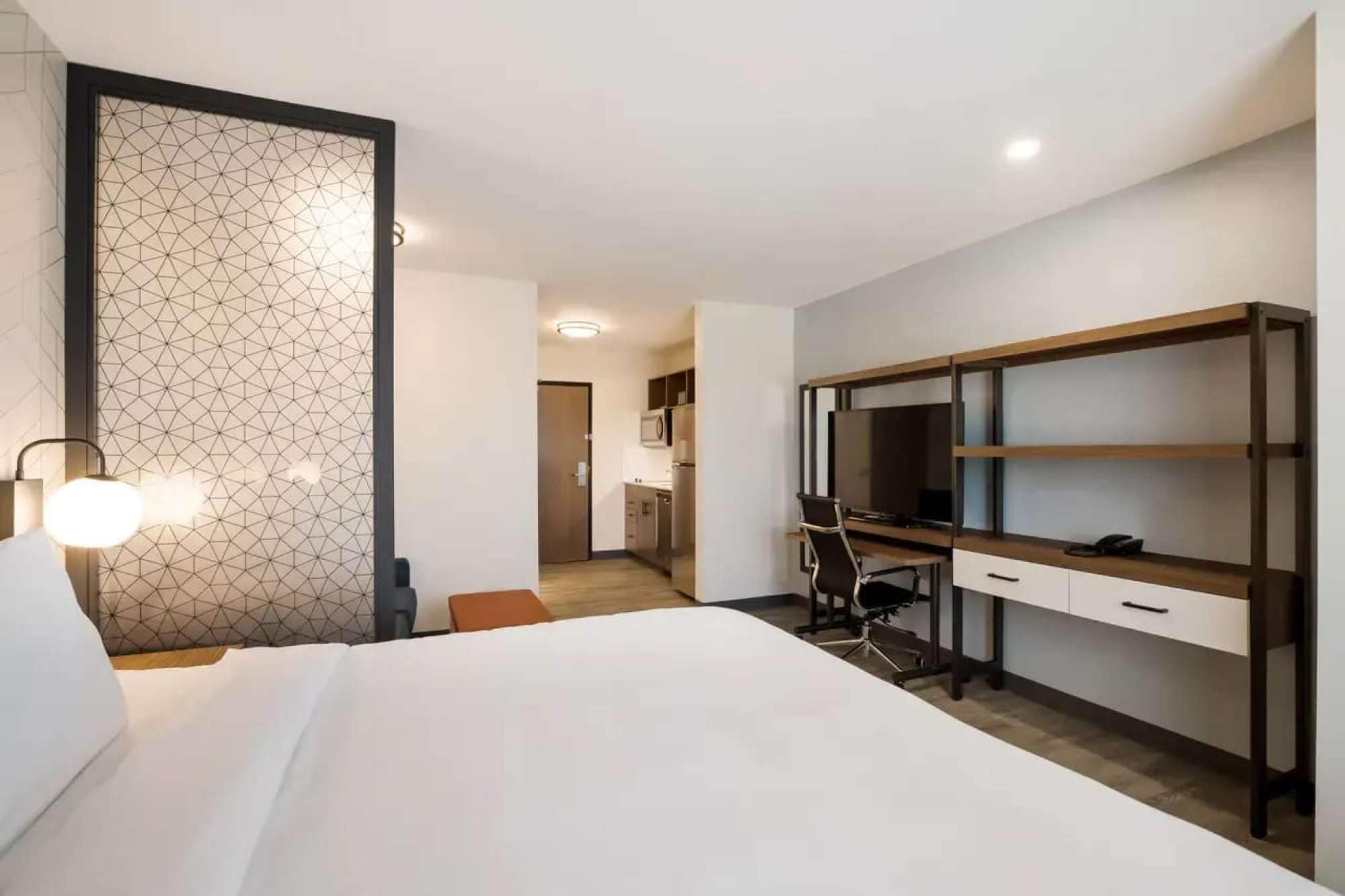 Une chambre d’hôtel avec un lit, un bureau et une chaise, un téléviseur et, au fond, une petite cuisine.