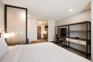 Een hotelkamer met een bed, een bureau met een stoel, een televisie en een kitchenette op de achtergrond.