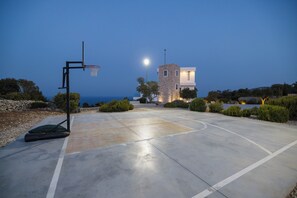 A basketball court with a hoop, a stone tower, and a modern building with a view of the sea. - The Luminous Villa: private estate, 4 sea-view suites, heated pool, sleeps 8 (Volimes)