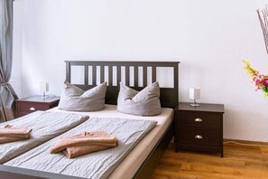 Een net opgemaakt bed met kussens en opgevouwen handdoeken, een nachtkastje met een lamp, en een aan de muur bevestigde vaas met bloemen.