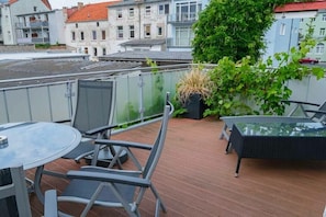 A rooftop terrace with a round glass table and chairs, a black wicker sofa, and a potted plant.