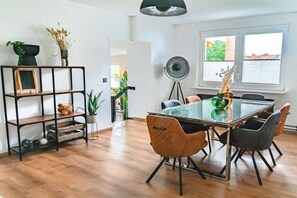 A dining area with a glass table, four chairs, a black metal shelf with decorative items, and a floor lamp.