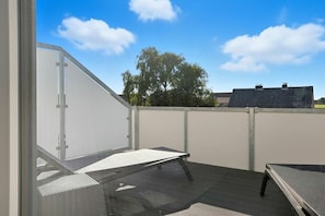 Varanda com porta de vidro, deck de madeira e vista para uma árvore e um edifício ao fundo.