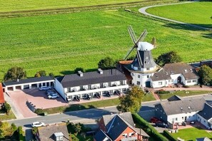Un molino de viento con un edificio blanco contiguo, un estacionamiento y un campo verde al fondo.