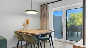 Uma área de refeições com mesa de madeira, cadeiras verdes e pretas, luminária pendente e vista para um pátio externo através de uma porta de vidro.