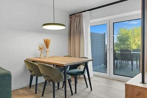 Un comedor con una mesa de madera, sillas verdes y negras, una lámpara colgante y vistas a un patio exterior a través de una puerta de vidrio.