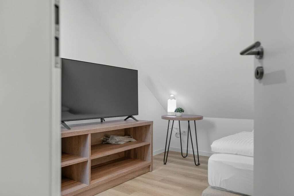 A modern room with a flat-screen TV on a wooden stand, a small round table with a plant, and a white bed.