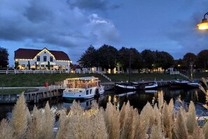 A riverside hotel with a boat docked in front, a well-lit building, and a calm water body. - Holiday apartment with 1 bedroom (Carolinensiel)