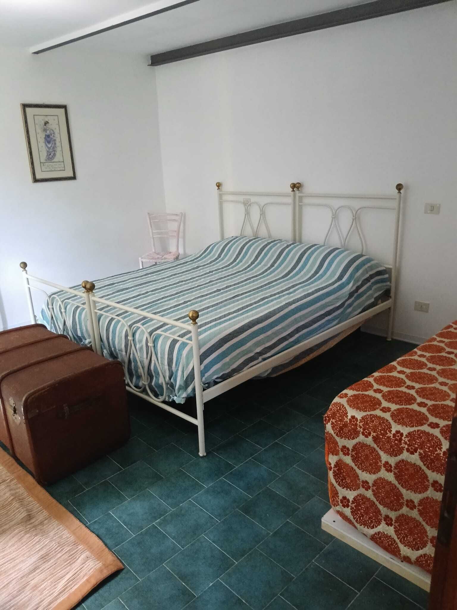 Un lit simple avec une couvre-lit à rayures, une chaise, une valise et un canapé à motifs dans une pièce dont le sol est recouvert de carreaux verts.