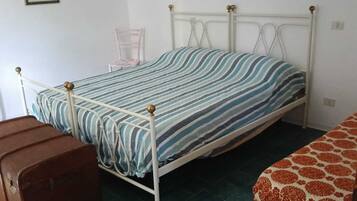 Un lit simple avec une couvre-lit à rayures, une chaise, une valise et un canapé à motifs dans une pièce dont le sol est recouvert de carreaux verts.