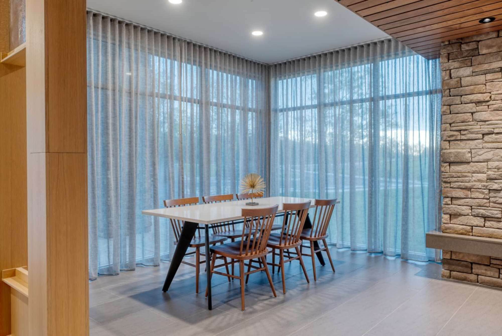 A dining area with a table and chairs, large windows with sheer curtains, and a stone accent wall.
