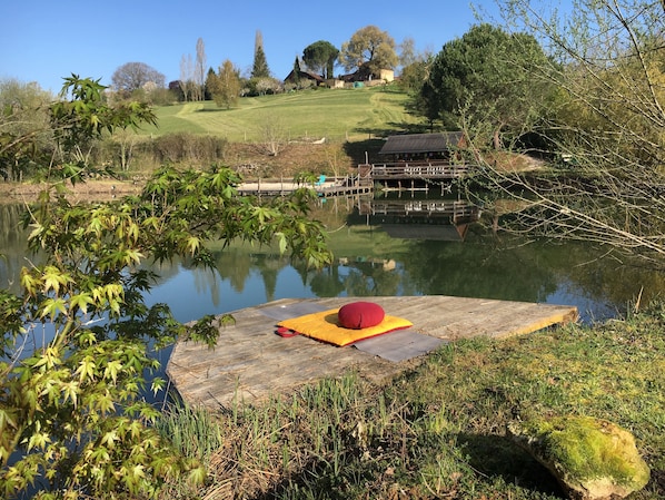 Un muelle de madera con un cojín rojo, un lago tranquilo y una colina cubierta de hierba con árboles al fondo.
