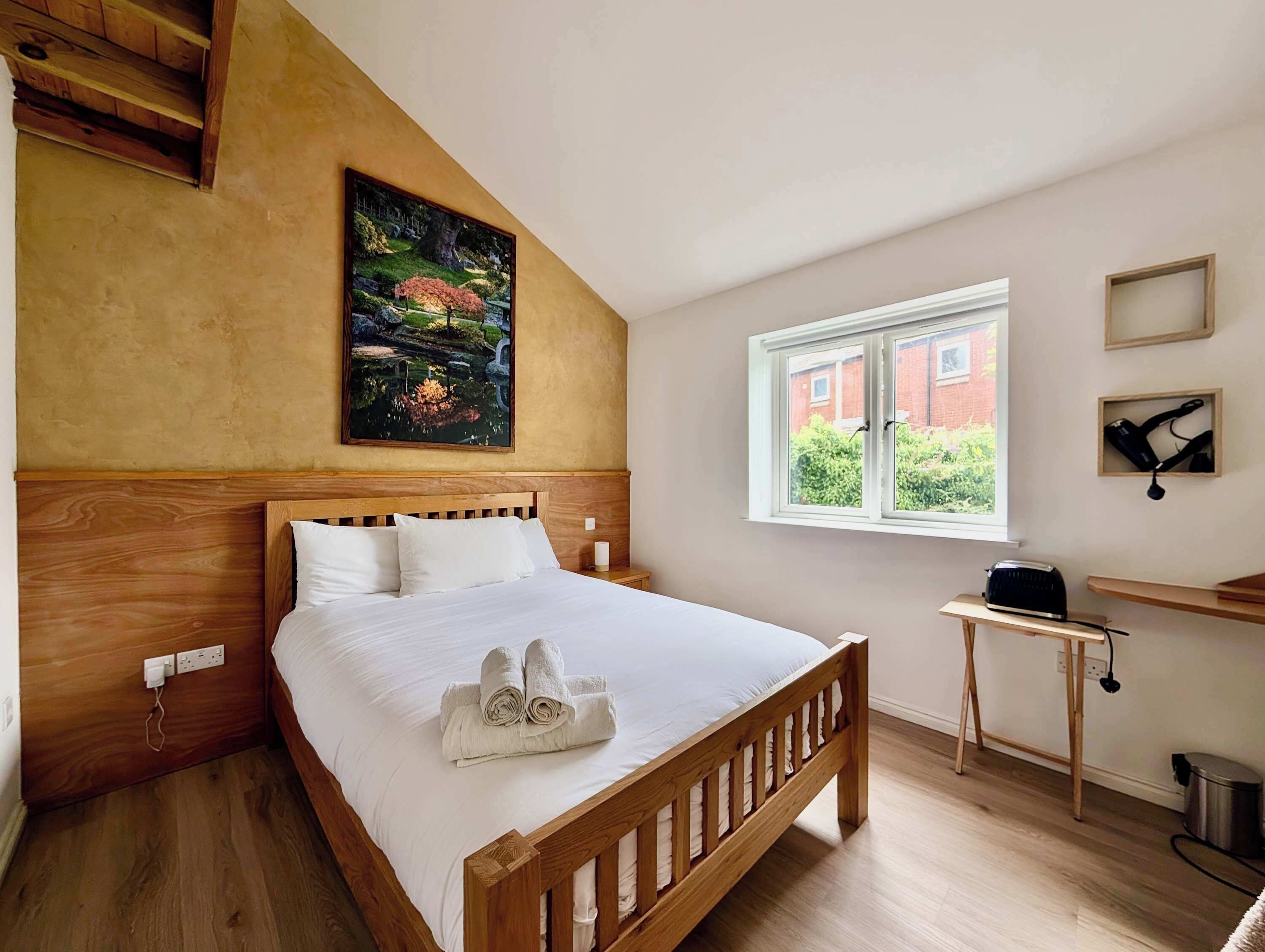 A bedroom with a wooden bed, a painting on the wall, a window with a view, and a small table with a toaster.