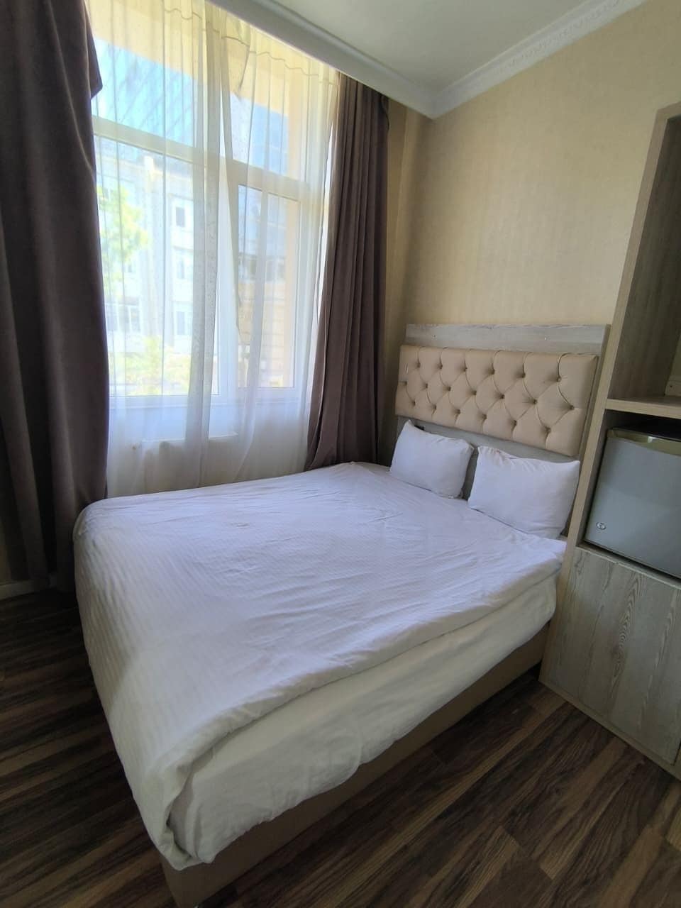 A hotel room with a large bed, a wooden floor, a window with curtains, and a small bedside table.