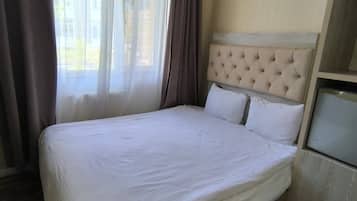 A hotel room with a large bed, a wooden floor, a window with curtains, and a small bedside table.