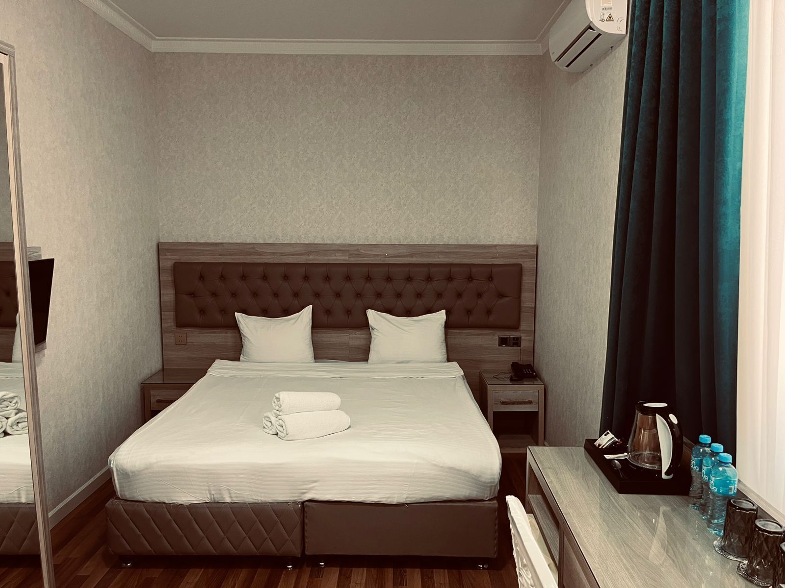 Una habitación de hotel con cama, mesitas de noche, un espejo, una ventana con cortinas y un aire acondicionado de pared.