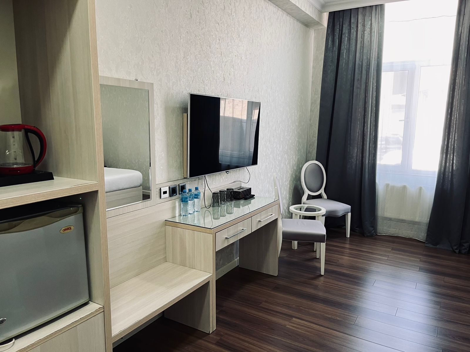 Habitación de hotel con cama, televisor, escritorio con espejo, refrigerador pequeño, silla y ventana con cortinas.