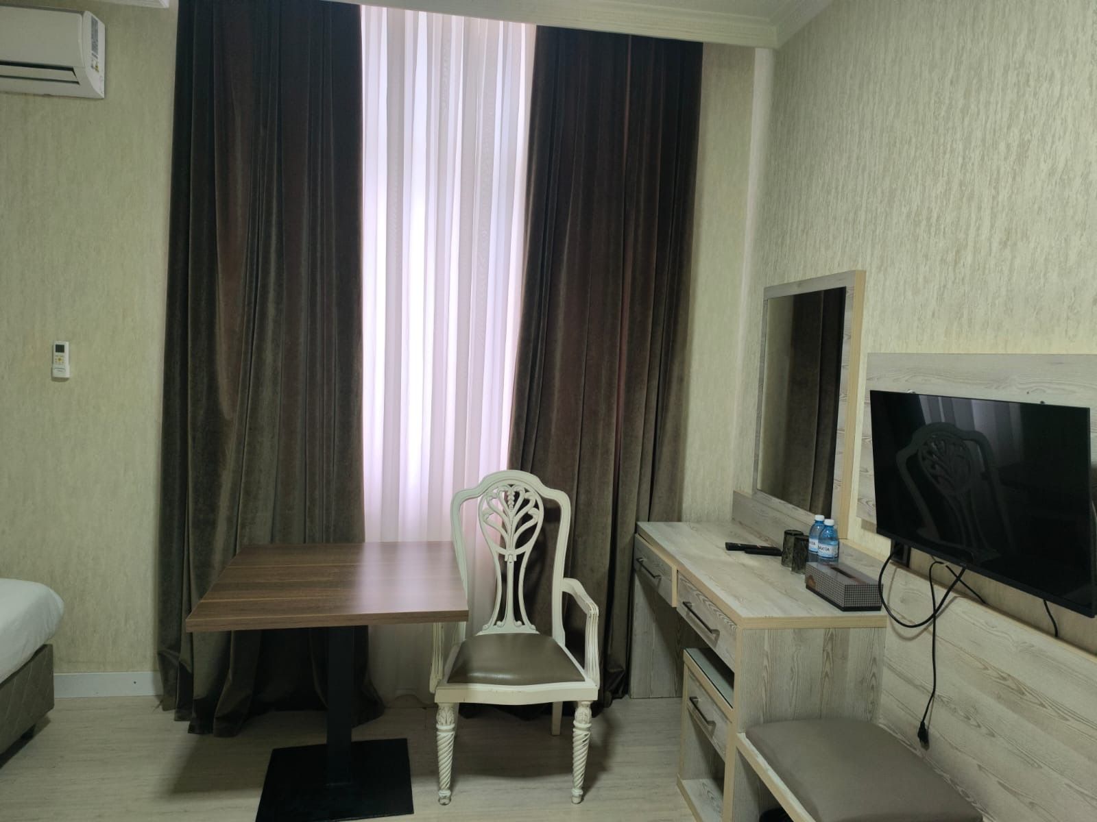 Una habitación de hotel con una cama, un escritorio con silla, un televisor y un ventanal con cortinas.
