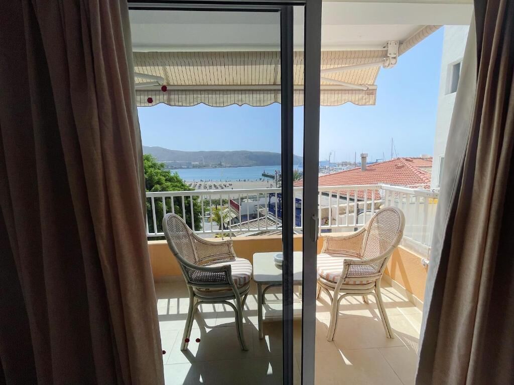 Varanda com cadeiras de vime e uma mesa, com vista para a área costeira, edifícios e céu claro.