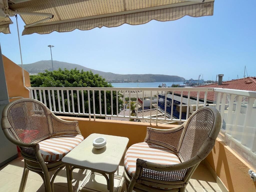 Un balcón con muebles de mimbre y una mesa, con vistas a una zona costera con edificios y una montaña al fondo.