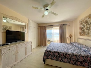 Habitación de hotel con cama, televisor, ventilador de techo, ventana grande con cortinas y vista al mar.