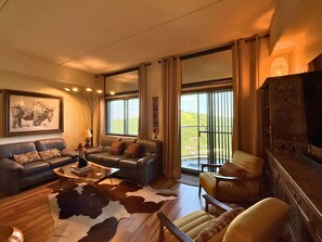 Una sala de estar con sofá, sillón y mesa de centro. Hay un ventanal con cortinas, una obra de arte colgada en la pared y una chimenea.