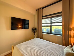 A bedroom with a bed, a wall-mounted TV, a standing fan, and a window with blinds. - Welcome to your mountain getaway! 2 bedroom condo is located on the 8th floor (Sugar Mountain)
