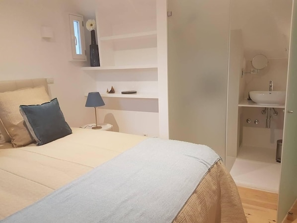 A bedroom with a bed, a bedside table, a lamp, a shelf with decorative items, and a bathroom with a sink and mirror. - Duplex near the Arch (Lisboa)