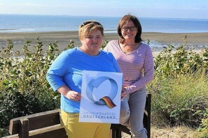 Deux personnes tenant une pancarte ornée d’un logo, debout à côté d’un banc en bois, avec une vue sur la mer.