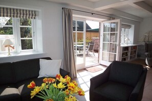 A living room with a sofa, a chair, a window with curtains, and a view of an outdoor patio. - De Gröne Eck 4 House 2 (Nieblum)