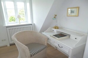 A room with a white desk, a wicker chair, a radiator, a window with a lamp, and a framed picture on the wall. - De Gröne Eck 4 House 2 (Nieblum)