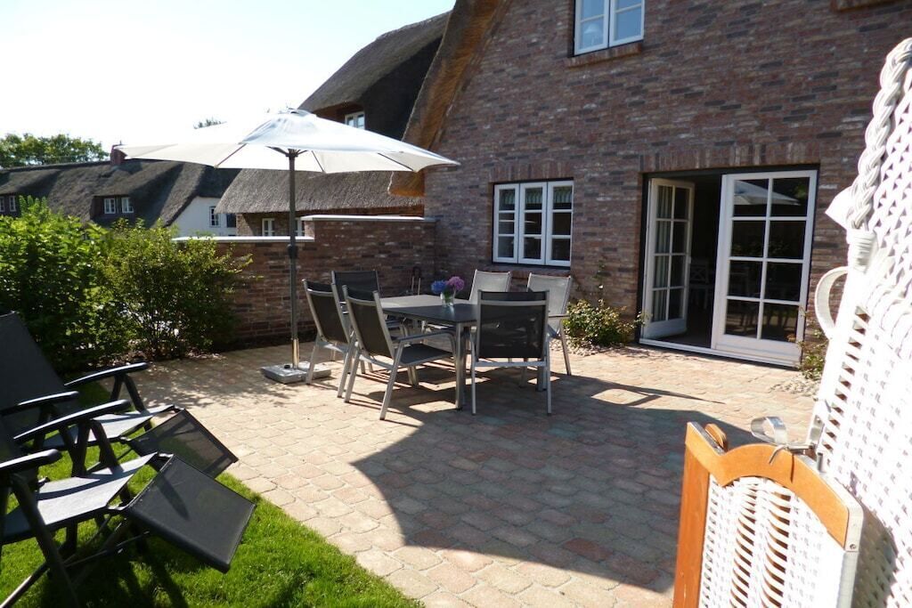 Een terras met een tafel, stoelen, een witte parasol, en een bakstenen gebouw met een raam en een deur.