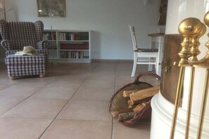 A living room with a checkered armchair, a white chair, a bookshelf, and a fireplace with a basket of wood. - De Gröne Eck 4 House 2 (Nieblum)