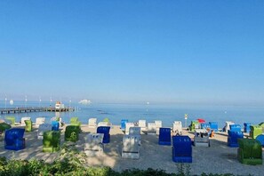 Ein Strand mit Reihen von nummerierten Liegestühlen und Sonnenschirmen, ein Steg im Hintergrund und das Meer.