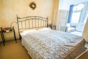 A bedroom with a metal bed frame, a patterned bedspread, a bedside table with a book, and a window with blinds. - Tower House Holiday Apartment 6 (Kühlungsborn)