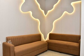 A corner room with a brown sofa, a wall with a unique light design, and a plain white wall. - ANJU VILLA (Varanasi)