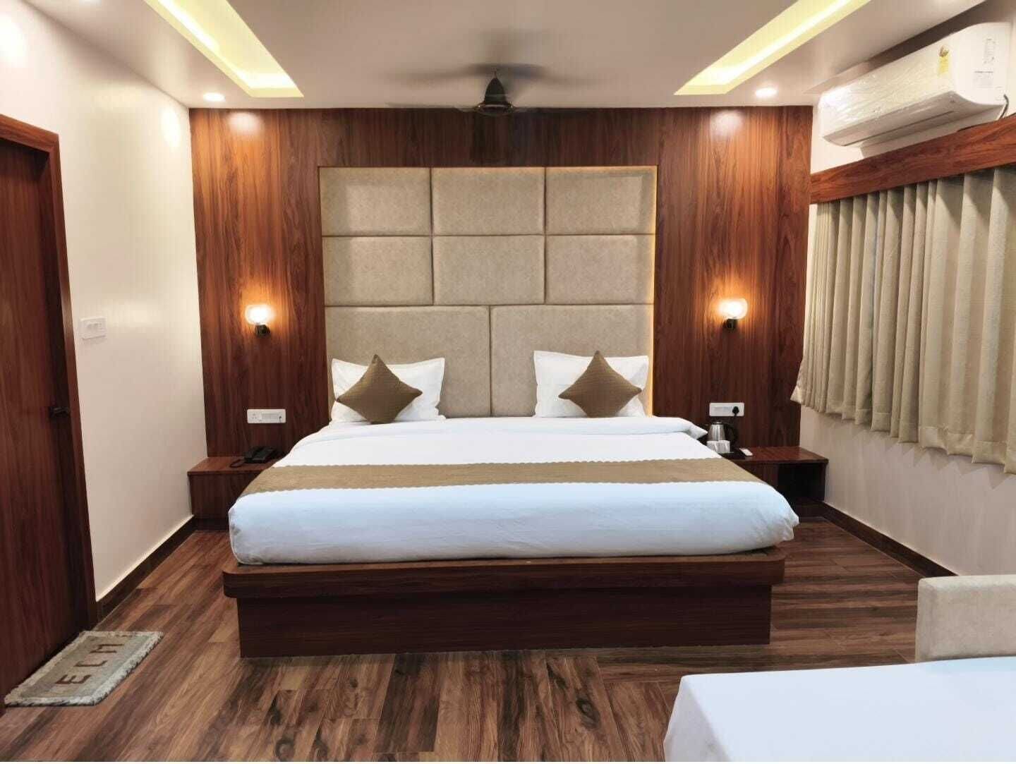 A hotel room with a large bed, wooden paneling, a headboard with a padded section, two bedside tables with lamps, and a window with curtains.