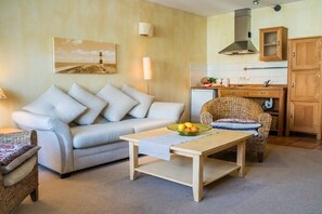 A cozy living room with a sofa, a dining table, and a wicker chair. A kitchen with wooden cabinets and a small refrigerator is visible in the background. - Tower House Holiday Apartment 5 (Kühlungsborn)