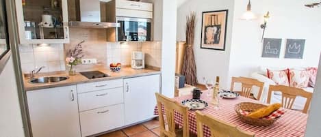 Cocina compacta con fregadero, estufa y microondas. Hay una mesa de comedor con sillas y mantel a cuadros rojo.