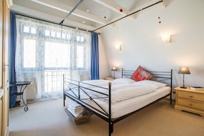 A bedroom with a metal bed frame, a bedside table with a lamp, a window with curtains, and a small table with a chair. - Tower House Holiday Apartment 9 (Kühlungsborn)